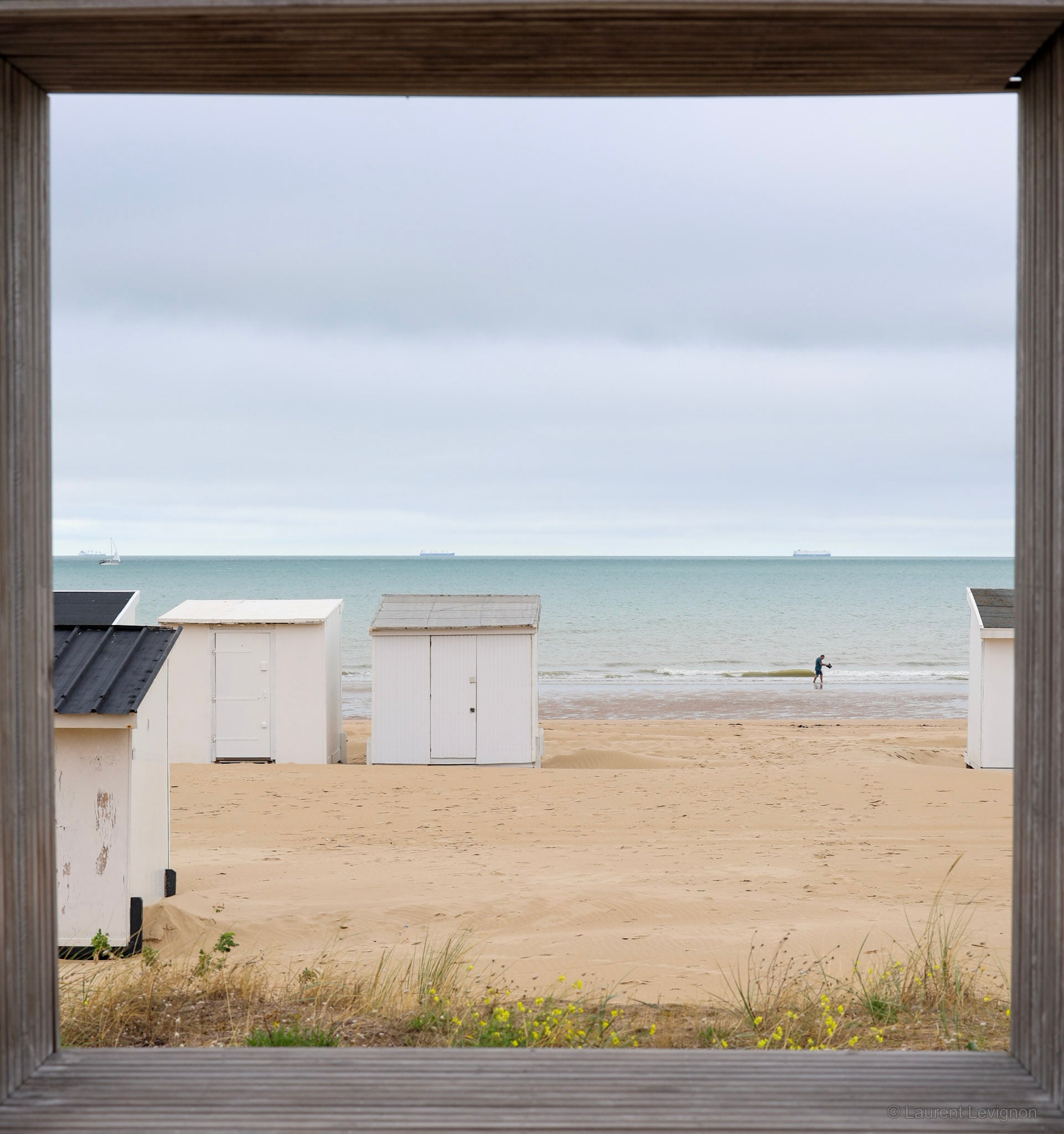 Vue depuis l'un des petits abris où l'on s'assoit pour manger face à la mer : les chalets de plage, une silhouette dans les vagues et, au large, deux cargos. Septembre 2026.