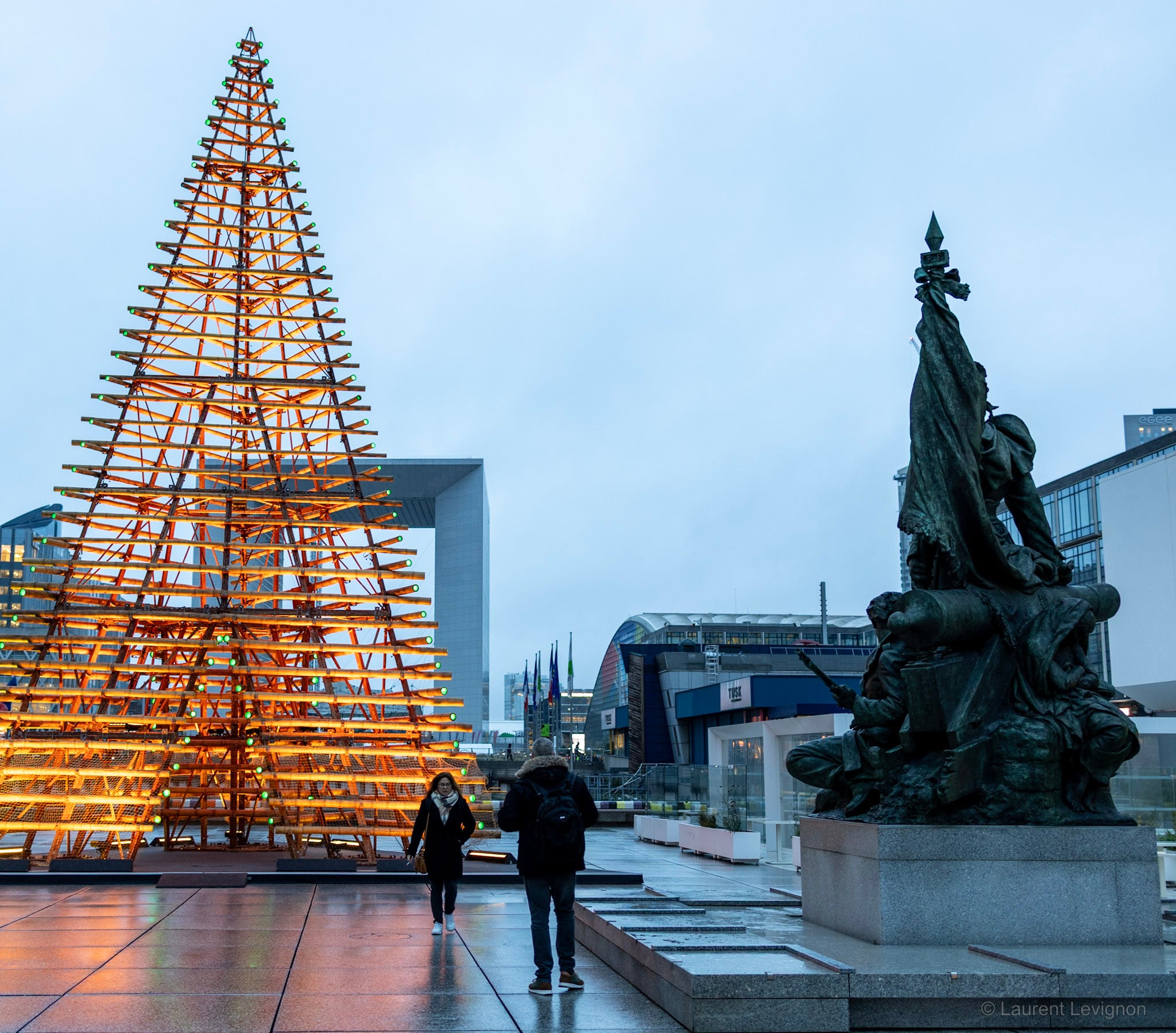 Le parvis en décembre 2023 : le sapin de bois, la Grande Arche prise dans sa silhouette, et à droite « La Défense de Paris » de Barrias, 1883 — le monument qui a donné son nom au quartier.